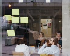 Teilnehmer*innen einer Kreativ-Werkstatt sitzen konzentriert in einem Workshop zusammen und halten ihre Arbeitsergebnisse auf Klebezetteln an einer Fensterscheibe fest. 